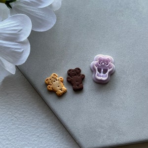 May include: Three cookie cutters in the shape of bears. The cutters are made of purple plastic and are on a gray surface. Two cookies are also shown, one brown and one light brown.