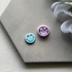 May include: Two cookie cutters in the shape of a smiley face with star eyes. One is light blue and the other is purple.