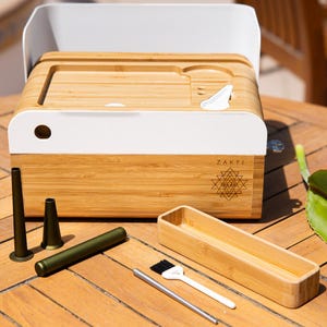 May include: A bamboo box with a white top and the word "ZAKTI" printed on the front. The box has a geometric design etched into the wood. Accessories include metal tubes, a brush, and a bamboo tray.
