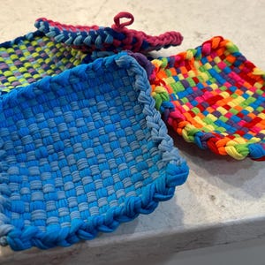 May include: Four colorful, woven pot holders in shades of blue, green, pink, yellow, orange, and red. Each pot holder is square-shaped with a braided edge and a loop for hanging. The pot holders are made from recycled fabric.