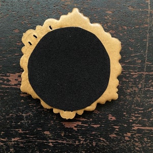 Op de afbeelding: Een goudkleurig, sierlijk frame met een zwarte cirkelvormige inzet. Het frame heeft een geschulpte rand en kleine gaatjes aan de bovenkant. Het object staat op een donkere, getextureerde houten ondergrond.