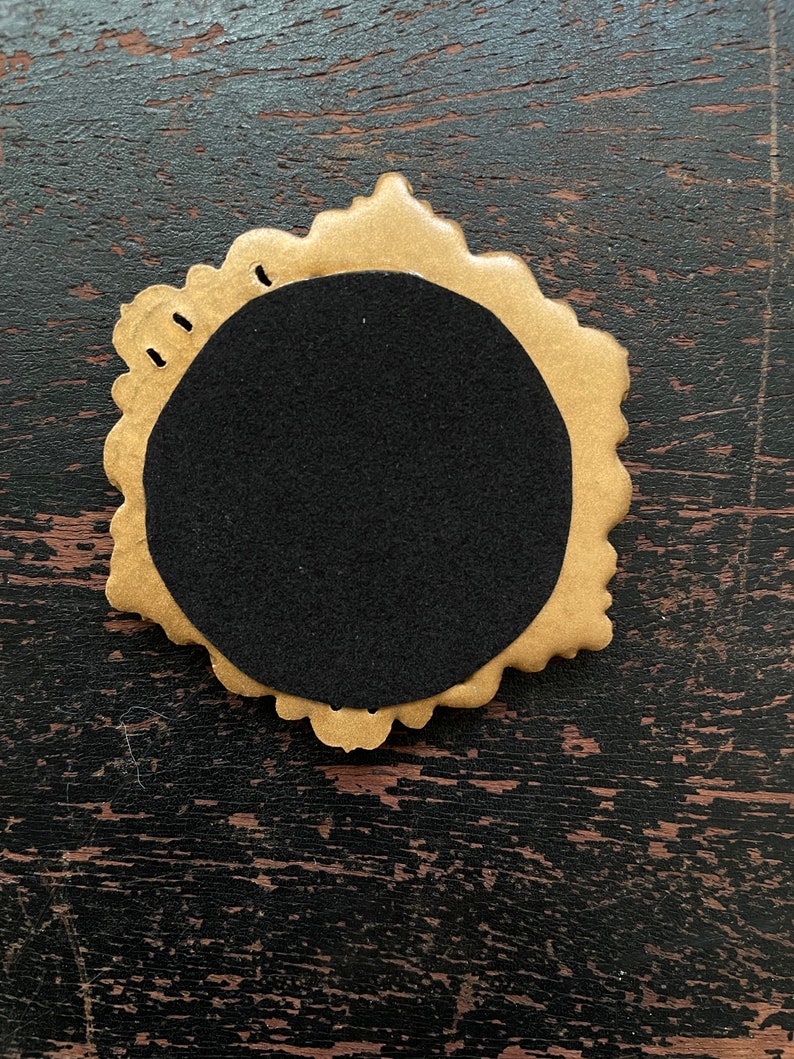 Op de afbeelding: Een goudkleurig, sierlijk frame met een zwarte cirkelvormige inzet. Het frame heeft een geschulpte rand en kleine gaatjes aan de bovenkant. Het object staat op een donkere, getextureerde houten ondergrond.