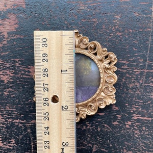 Op de afbeelding: Ovaal fotolijstje in goudkleur met een kleine landschapsafbeelding. Het frame is ongeveer 7,6 cm hoog, gemeten met een houten liniaal. Het frame heeft een gedetailleerd, krullend ontwerp.