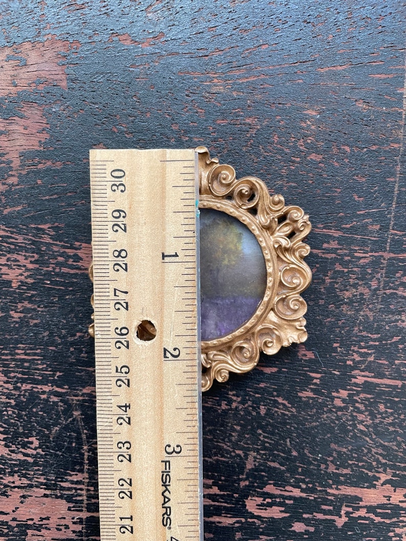 Op de afbeelding: Ovaal fotolijstje in goudkleur met een kleine landschapsafbeelding. Het frame is ongeveer 7,6 cm hoog, gemeten met een houten liniaal. Het frame heeft een gedetailleerd, krullend ontwerp.