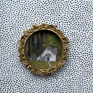 May include: A gold-toned oval frame with a miniature painting of a white cottage in a forest setting. The painting is set against a white background with black polka dots.
