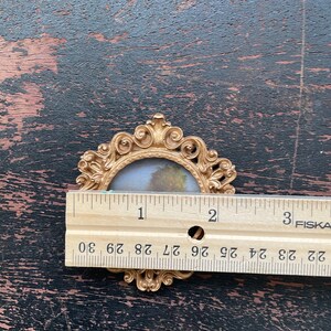 Op de afbeelding: Een kleine, sierlijke goudkleurige fotolijst met een landschapsschilderij erin. Het frame is ongeveer 5 cm breed. Het frame heeft een decoratief, scrollachtig ontwerp rond de randen. De achtergrond is een donker, verweerd houten oppervlak.