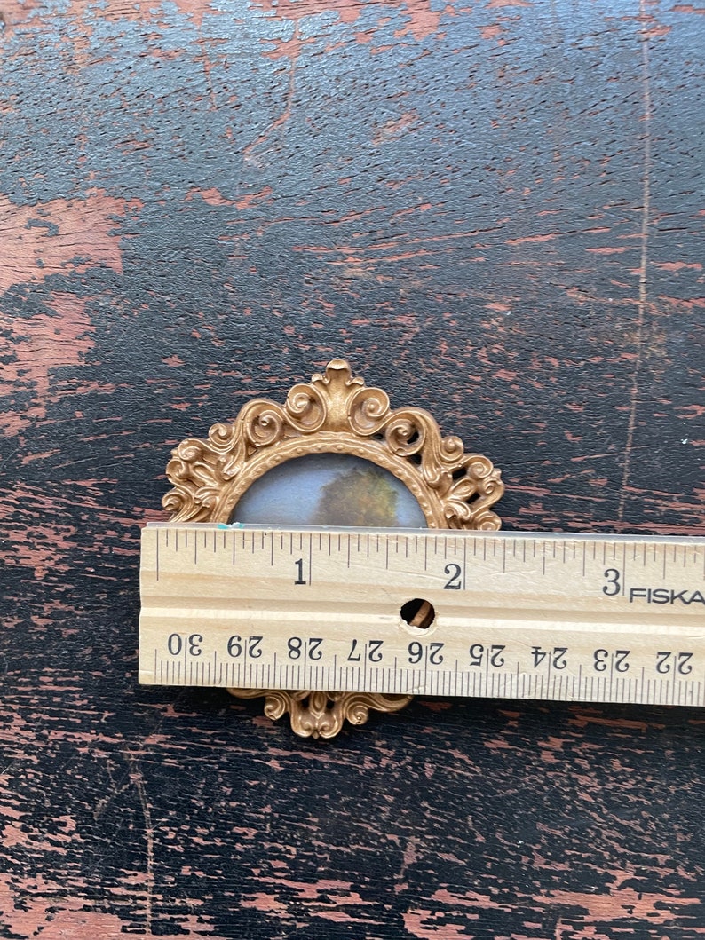 Op de afbeelding: Een kleine, sierlijke goudkleurige fotolijst met een landschapsschilderij erin. Het frame is ongeveer 5 cm breed. Het frame heeft een decoratief, scrollachtig ontwerp rond de randen. De achtergrond is een donker, verweerd houten oppervlak.
