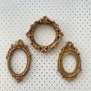 May include: Three ornate, gold-colored picture frames of varying shapes: oval, round, and diamond. The frames are empty, with intricate detailing. They are arranged against a white background with gold polka dots.