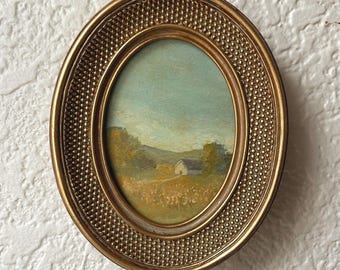May include: Oval-shaped framed landscape painting. The painting depicts a small white house in a field, with trees and hills in the background. The frame is gold-colored with a textured, dotted pattern.