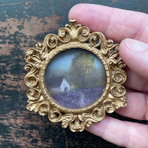 Op de afbeelding: Omsloten gouden miniatuurframe met een rond landschapsschilderij. De schildering toont een wit gebouw, bomen en een veld met paarse bloemen. Het frame heeft een gedetailleerd, scrollend ontwerp.