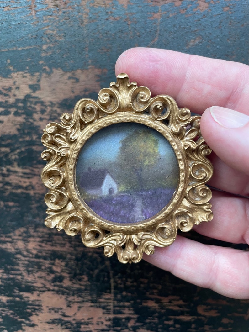 Op de afbeelding: Omsloten gouden miniatuurframe met een rond landschapsschilderij. De schildering toont een wit gebouw, bomen en een veld met paarse bloemen. Het frame heeft een gedetailleerd, scrollend ontwerp.