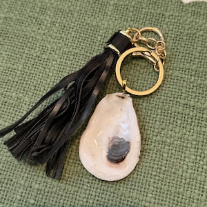 May include: A keychain featuring a polished white oyster shell with a dark gray center, attached to a gold-tone metal ring and a black faux leather tassel. The hardware includes a gold-tone jump ring and a lobster clasp for secure attachment.