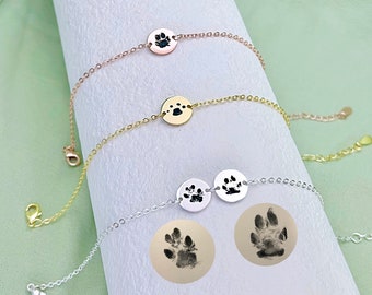 May include: Three silver bracelets with paw print charms. The bracelets are layered on a white surface. The charms are round and have a black paw print design.