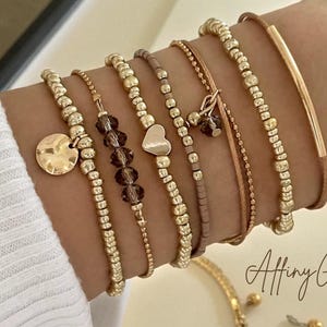 Può includere: Una collezione di braccialetti color oro con vari design. Alcuni braccialetti presentano piccole perline, un ciondolo a forma di cuore e un ciondolo a disco. Altri braccialetti sono fatti di un cordoncino sottile. I braccialetti sono impilati su un polso.