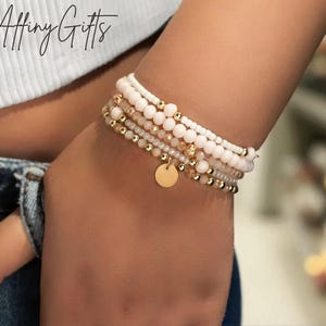 May include: A stack of beaded bracelets in shades of white, pink, and gold. The bracelets feature various bead sizes and textures, including small gold beads and a gold disc charm. The bracelets are stacked on a wrist, with the brand name "Affiny Gifts" in the top left corner.