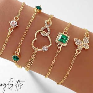 Peut inclure: Bracelets dorés avec divers motifs, dont un cœur, un papillon et une pierre carrée vert émeraude. Les bracelets sont ornés de pierres précieuses claires et vert émeraude. Le texte "Affiny Gifts" est en bas.