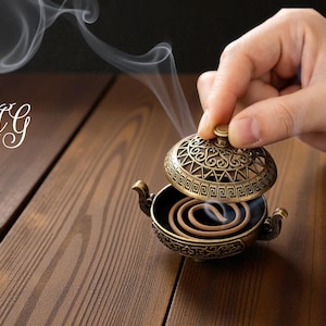 May include: A decorative bronze incense burner with an open lid, releasing wisps of smoke. The burner features intricate patterns and holds a coiled incense stick. The initials "AG" are in the upper left corner.