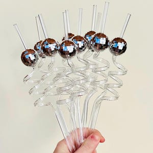 May include: A collection of clear plastic cocktail stirrers, each featuring a small, mirrored disco ball. The stirrers have a swirled design near the ball and are held in a hand against a plain background. Ideal for adding a touch of fun to any drink.