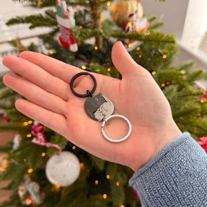 May include: A keychain with a circular pendant showing two intertwined cat silhouettes, one grey and one white. The keychain has a black ring and a silver ring. The background includes a Christmas tree with decorations.