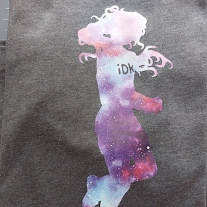 May include: A gray t-shirt with a galaxy print silhouette of a person jumping. The silhouette has the text "idk" printed on it.
