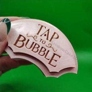 May include: A light pink, semi-circular stamp with the words "TAP TO BUBBLE" in a brown, embossed font. The stamp has a decorative border and is held against a green background. The text is in a stylized font.