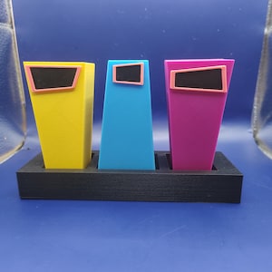 Könnte beinhalten: Drei aufrechte, konisch zulaufende, trapezförmige Behälter in leuchtendem Gelb, Blau und Magenta, die jeweils mit einem kleinen, schwarz gefüllten und rosa umrandeten Rechteck versehen sind. Die Behälter stehen in einem schwarzen Sockel.