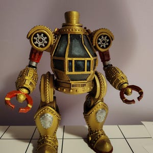 May include: A golden-colored steampunk robot figurine with intricate details. The robot has a cylindrical body with a glass-like compartment, gear-shaped shoulder pads, and articulated arms ending in red claw-like hands. The legs are also gold with red accents.