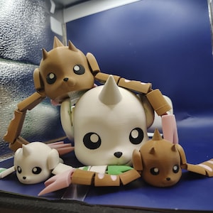 May include: Three articulated figures with large heads and pointed horns. One large white figure is centered, flanked by two smaller figures, one brown and one white. Each figure features large black eyes, small noses, and segmented limbs.