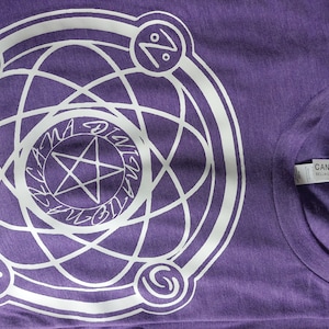 Może przedstawiać: Fioletowy T-shirt z białym nadrukiem w kształcie koła z pięcioramienną gwiazdą w środku i napisem "Digital Alchemy" wokół niej. Wokół koła znajdują się cztery symbole.