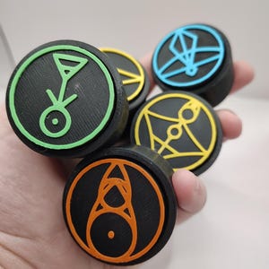 Op de afbeelding: Een verzameling zwarte, cirkelvormige objecten met kleurrijke geometrische ontwerpen in groen, geel, oranje en blauw. De objecten worden in een hand gehouden en de ontwerpen staan tegen een zwarte achtergrond.