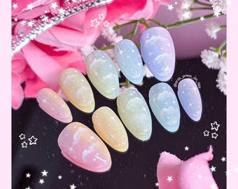Puede incluir: Un conjunto de diez uñas postizas en colores pastel. Cada uña presenta un diseño de nube con detalles brillantes. Los colores incluyen rosa, amarillo, verde y azul. Las uñas están dispuestas sobre una superficie reflectante con decoraciones florales y estrellas.