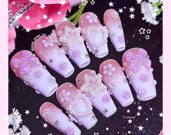 Può includere: Un set di dieci unghie finte con un design sfumato rosa e bianco. Ogni unghia presenta elementi decorativi come fiori, perle e fiocchi. Alcune unghie hanno un design a forma di cuore. Le unghie sono disposte su una superficie riflettente.