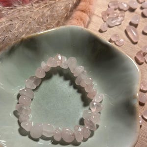 Könnte beinhalten: Ein Armband aus Rosenquarz-Chip-Perlen liegt in einer hellgrünen Keramikschale. Das Armband besteht aus kleinen, polierten, hellrosa Steinen. Zusätzliche Rosenquarz-Chips sind um die Schale herum verstreut. Trockenblumen sind im Hintergrund.