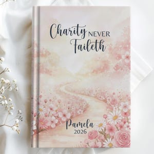 May include: A hardcover journal with a watercolor design. The cover features the text "Charity Never Faileth" and "Pamela 2026". The artwork depicts a path winding through a field of pink flowers and a soft, dreamy sky.