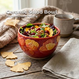 Puede incluir: Un cuenco de cerámica rojo con diseños florales amarillos pintados a mano, lleno de ensalada, nueces y arándanos. El texto "Ceramic Salad Bowl" y "Hand painted Pottery" es visible. Una taza y una servilleta están en el fondo.