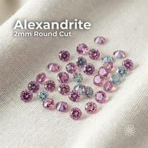 May include: A collection of 2mm round-cut alexandrite gemstones in shades of pink and blue-green, scattered on a white fabric. The gemstones are faceted, reflecting light. The text "Alexandrite 2mm Round Cut" is displayed at the top.