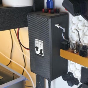 May include: A black electrical component with a carbon fiber texture. It features a circuit breaker and various wires and connectors. The component is mounted on a white and yellow surface, with additional black and red components visible.