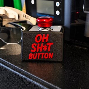 May include: A black square-shaped device featuring a large, circular, metallic red emergency-style button on top. The front face of the black housing displays the text "OH SH*T BUTTON" in bold, raised red lettering with a textured finish.