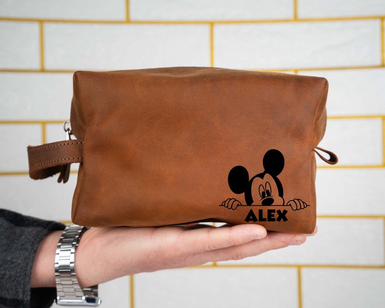 May include: Brown leather toiletry bag with a black Mickey Mouse design and the name "ALEX" printed on the front.