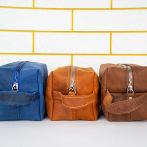 May include: Three leather toiletry bags in different colors: blue, brown, and orange. Each bag has a zipper closure and a leather handle.