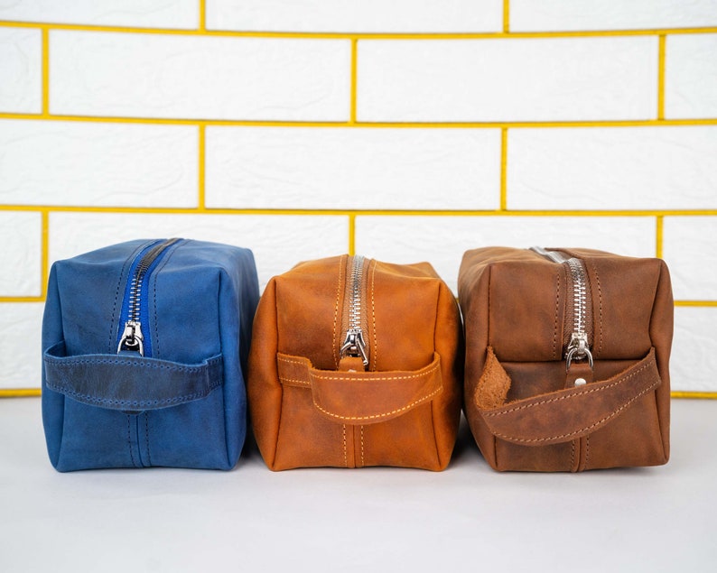 May include: Three leather toiletry bags in different colors: blue, brown, and orange. Each bag has a zipper closure and a leather handle.