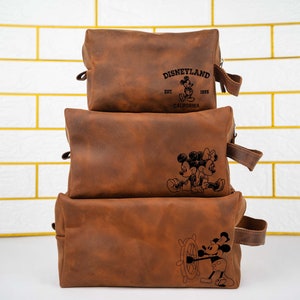 May include: Three brown leather travel bags stacked on top of each other. The top bag has the text "DISNEYLAND EST. 1955 CALIFORNIA" and a Mickey Mouse and Minnie Mouse design. The middle bag has a Mickey Mouse and Minnie Mouse design. The bottom bag has a Mickey Mouse design with a ship's wheel.