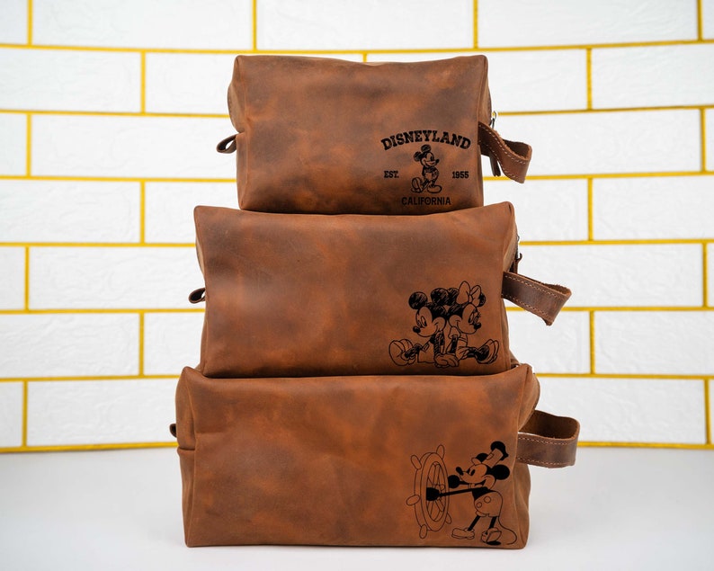May include: Three brown leather travel bags stacked on top of each other. The top bag has the text "DISNEYLAND EST. 1955 CALIFORNIA" and a Mickey Mouse and Minnie Mouse design. The middle bag has a Mickey Mouse and Minnie Mouse design. The bottom bag has a Mickey Mouse design with a ship's wheel.