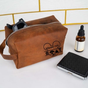 May include: Brown leather toiletry bag with a zipper closure and a black handle. The bag has a Mickey Mouse design with the text "Disney Trip 2024" and icons of a castle, a tree, and a roller coaster.