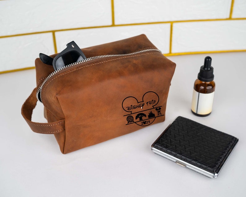May include: Brown leather toiletry bag with a zipper closure and a black handle. The bag has a Mickey Mouse design with the text "Disney Trip 2024" and icons of a castle, a tree, and a roller coaster.