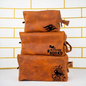 May include: Three brown leather travel bags stacked on top of each other. The top bag has a Mickey Mouse head and a ship's wheel design. The middle bag has the text "#Disney Squad". The bottom bag has a ship and the text "Let's Cruise".