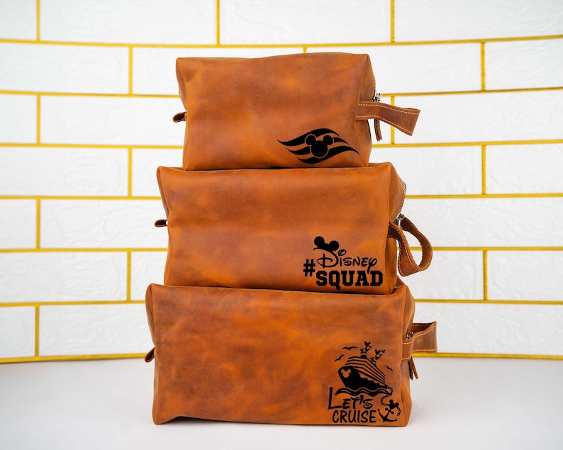 May include: Three brown leather travel bags stacked on top of each other. The top bag has a Mickey Mouse head and a ship's wheel design. The middle bag has the text "#Disney Squad". The bottom bag has a ship and the text "Let's Cruise".