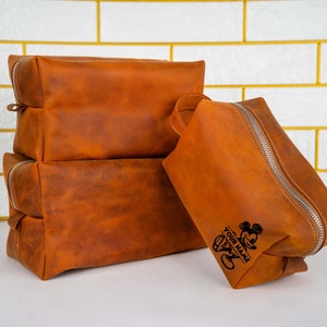 May include: A set of three brown leather travel bags with a zipper closure. The largest bag has a Mickey Mouse design and the text "YOUR NAME" engraved on it.