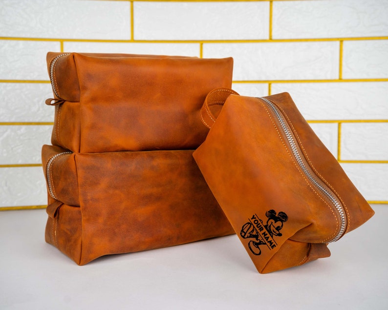 May include: A set of three brown leather travel bags with a zipper closure. The largest bag has a Mickey Mouse design and the text "YOUR NAME" engraved on it.