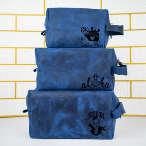 May include: Three blue toiletry bags stacked on top of each other. The top bag has the text "On Cruise Mode" with a cruise ship and Mickey Mouse ears. The middle bag has the text "Aloha" with a pineapple and Mickey Mouse ears. The bottom bag has the text "Oregon" with a cartoon duck wearing a football jersey.